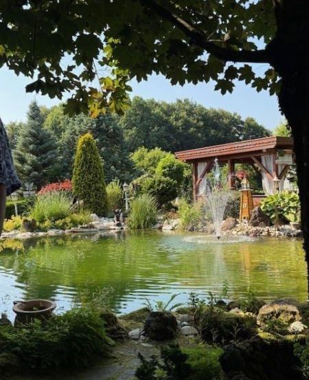 Ein malerischer Garten mit einem Teich und einem Springbrunnen. Der Teich ist von üppigem Grün und Felsen umgeben. Ein Pavillon mit Dach steht in der Nähe des Wassers, und es gibt Bäume und Sträucher im Hintergrund.