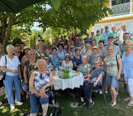 Eine große Gruppe von Menschen, wahrscheinlich Familie oder Freunde, stehen und sitzen um einen Tisch in einem Garten, mit einem Gebäude und Bäumen im Hintergrund.