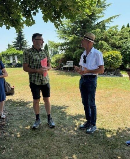 Zwei Männer unterhalten sich in einem Park mit üppiger Grünfläche und klarem Himmel. Ein Mann trägt einen Hut und eine Sonnenbrille, während der andere eine rosa Mappe in der Hand hält.