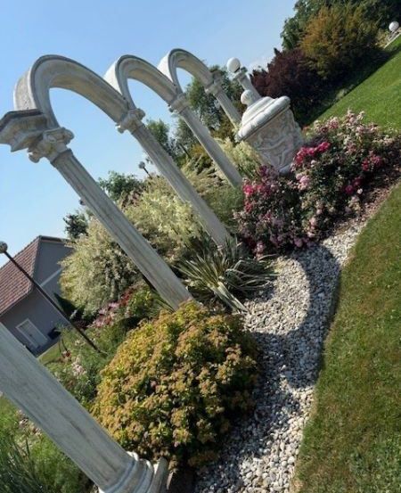 Ein Garten mit gebogenen Steinstrukturen, umgeben von bunten Blumen, Büschen und Bäumen, mit einem Haus im Hintergrund.