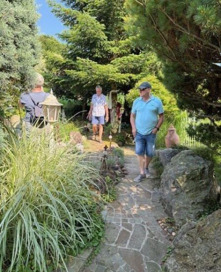 Drei Männer gehen auf einem Steinweg in einem Garten, umgeben von hohen Bäumen und Pflanzen. Ein Mann trägt einen Rucksack.