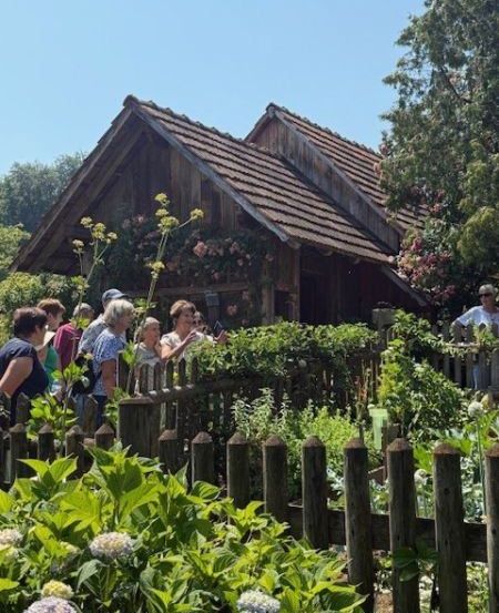 Eine Gruppe von Menschen steht vor einem Holzhaus mit Garten und betrachtet die Blumen.