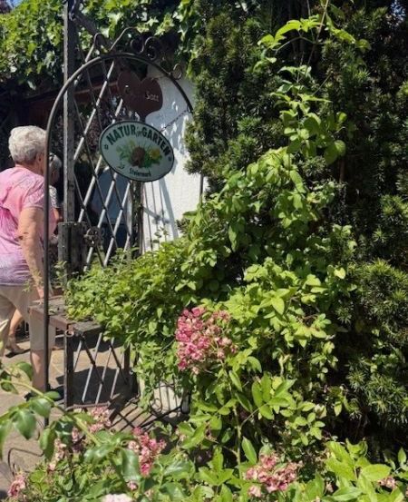 Eine Frau geht auf einen Garteneingang mit einem Schild zu, das Naturgarten sagt. Sie trägt ein rosa Shirt und beige Hose. Der Garten ist von rosa Blumen, grünen Blättern und einem Metalltor umgeben.