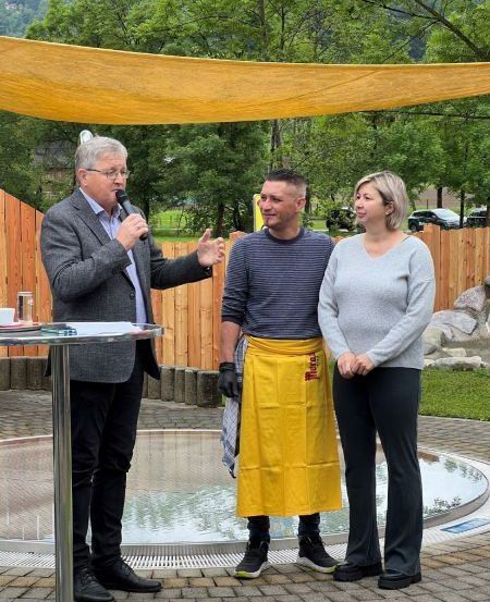 Ein Mann spricht in ein Mikrofon, während zwei andere neben ihm vor einem kleinen Pool stehen. Sie befinden sich in einem Garten mit einem Holzzaun und einem Baldachin.