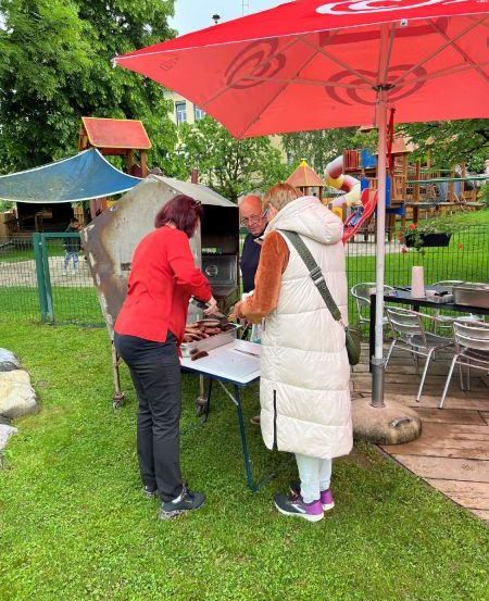 Zwei Frauen bereiten Essen an einem Tisch unter einem roten Schirm in einem Park zu. Eine weitere Person steht in der Nähe. Es gibt Tische und Stühle, einen Spielplatz und Bäume im Hintergrund.