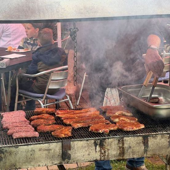 Eine Person grillt verschiedene Fleischsorten auf einem Grill, während andere am Tisch essen. Ein Kind beobachtet den Grillvorgang.