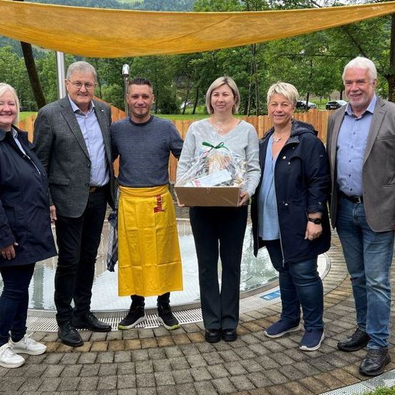 Fünf Menschen stehen draußen neben einem Brunnen und lächeln für ein Foto. Eine Frau hält eine Box mit einem Geschenk, und ein Mann trägt eine gelbe Schürze. Hinter ihnen ist ein gelber Baldachin über einem Holzzaun. Bäume und Berge sind im Hintergrund.