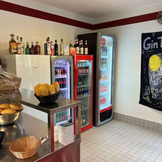 Eine Küche mit einer Theke hat verschiedene Brote, Getränke und Flaschen. Ein Schwarzes Brett mit den Worten 'Gin Tonic' und einer Zitronenillustration ist an der Wand befestigt.