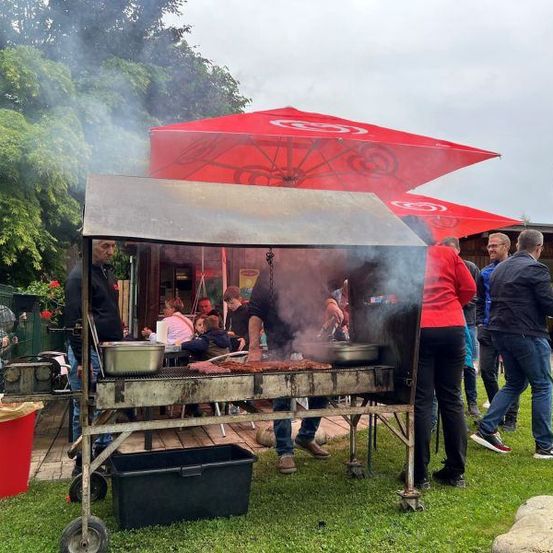 Eine Gruppe von Menschen grillt Essen auf einem Wagen unter roten Sonnenschirmen. Ein Mülleimer steht in der Nähe. Menschen gehen herum und schauen zu.