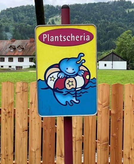 Ein Schild mit einer Cartoon-Figur, die in einem Pool schwimmt, steht vor einem Holzzaun. Im Hintergrund ist ein Rasenfeld mit Häusern und Bäumen zu sehen.