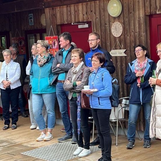 Eine Gruppe von Menschen steht in einem Raum mit Holzwänden, einige halten Getränke. Eine Person mit einer Tasse steht in der Mitte, und eine andere Person hält eine Flasche. Zwei Frauen stehen vor der Wand.