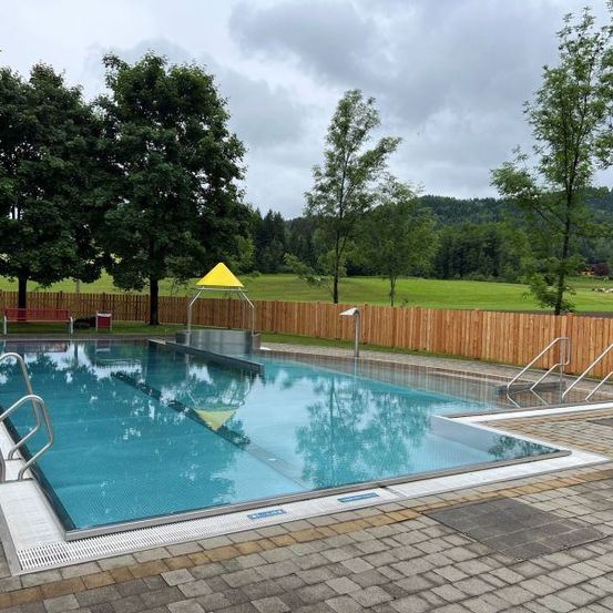Ein Freibad mit gelbem Baldachin und Handläufen ist von einem Holzzaun umgeben. Bäume und eine grüne Wiese sind im Hintergrund sichtbar.