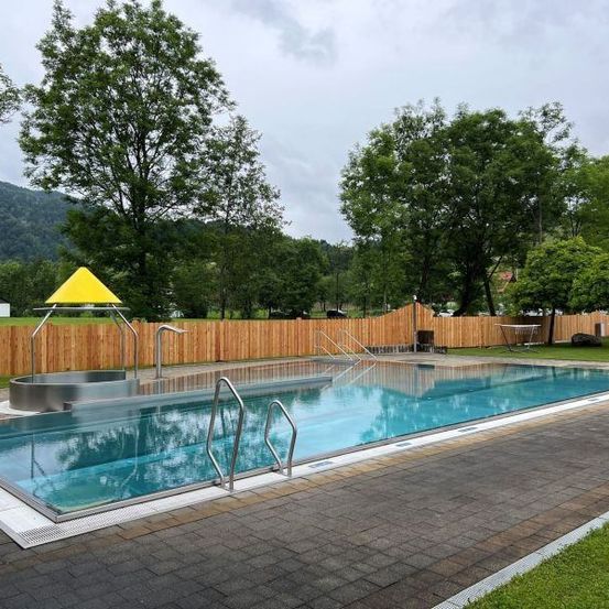 Ein Freibad mit gelbem Dach ist mit einem Holzzaun eingezäunt. Es gibt Bäume und Gras in der Umgebung. In der Ferne befindet sich ein Berg.