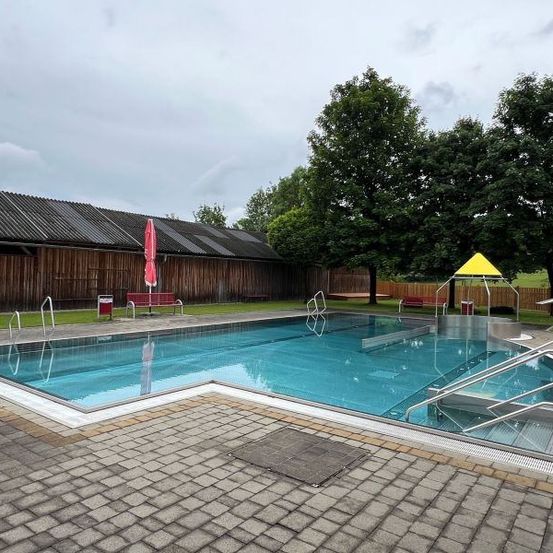 Ein Freibad mit gefliestem Boden und Handläufen. Es gibt einen geschlossenen roten Schirm und ein gelbes dreieckiges Objekt auf der rechten Seite. Hinter dem Pool steht ein Holzgebäude mit einem Metalldach. Im Hintergrund befinden sich Bäume und eine Rasenfläche.