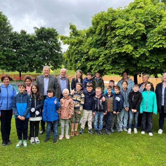 Eine Gruppe von Menschen, darunter Kinder und Erwachsene, posiert für ein Foto in einem Park mit Bäumen und einem Holzzaun.