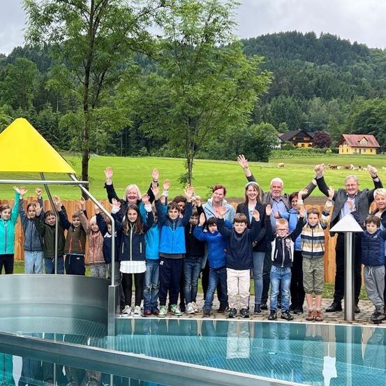 Eine Gruppe von Kindern und Erwachsenen posiert für ein Foto vor einem Schwimmbecken, mit einem gelben Baldachin und einer malerischen Aussicht auf Berge und ein Haus im Hintergrund.