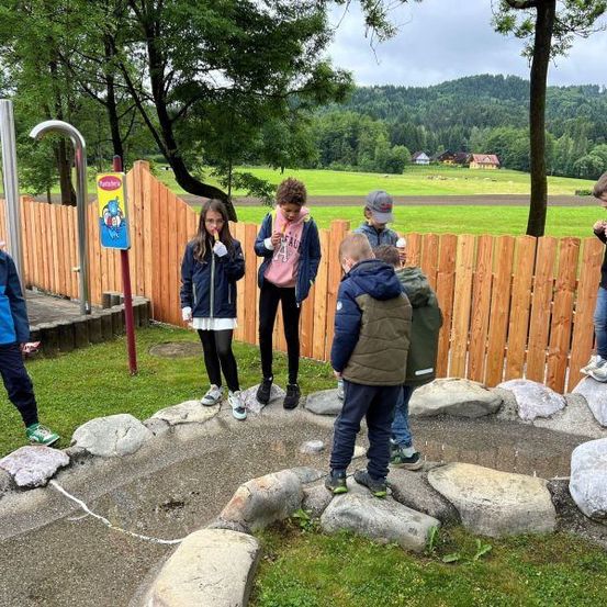 Eine Gruppe von Kindern steht auf Felsen in einem Park. Sie tragen Jacken und Sneaker. Hinter ihnen steht ein Holzzaun. In der Ferne befinden sich Häuser und Bäume.