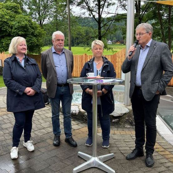 Vier Personen stehen in der Nähe eines Pools im Freien, eine hält ein Mikrofon. Zwei Frauen und zwei Männer, alle in Jacken gekleidet, scheinen bei einer Veranstaltung im Freien zu sprechen. Hinter ihnen befindet sich ein Holzzaun mit Bäumen und Bergen im Hintergrund.