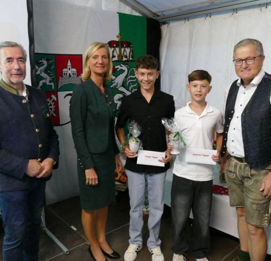 Fünf Menschen stehen in einer Reihe, lächeln, halten Tassen und feiern möglicherweise ein Ereignis. Sie stehen vor einem weißen Hintergrund mit einer Flagge. Der Mann auf der linken Seite trägt einen blauen Mantel, und der Mann auf der rechten Seite trägt ein weißes Hemd und eine schwarze Weste.