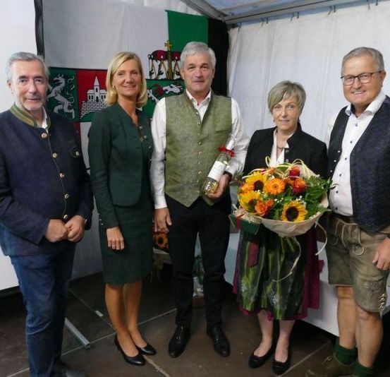 Fünf Personen stehen eng beieinander, möglicherweise bei einer Veranstaltung. Eine Frau hält einen Blumenstrauß. Der Mann in der Mitte hält eine Flasche.