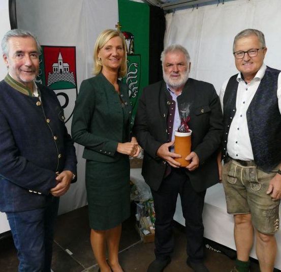 Vier Personen stehen in einem Zelt und lächeln. Ein Mann hält eine Trophäe. Eine Frau in einem grünen Kleid und ein Mann in einem schwarzen Anzug sind in der Nähe. Eine Fahne befindet sich hinter ihnen.
