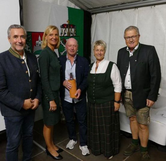 Fünf Menschen stehen lächelnd in einem Zelt und posieren für ein Foto. Ein Mann hält einen Pokal in der Hand, und hinter ihnen steht ein Tisch mit einer grünen Flasche.