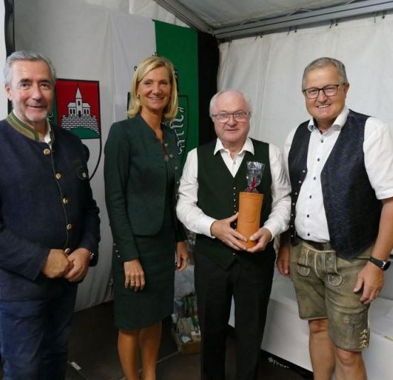Vier Personen stehen in einem Zelt. Ein Mann auf der linken Seite lächelt und trägt einen dunkelblauen Mantel. Eine Frau in der Mitte hält eine Trophäe in ihrer rechten Hand. Ein Mann auf der rechten Seite trägt eine Uhr und eine Weste. Hinter ihnen steht ein weißer Tisch mit einigen Flaschen.