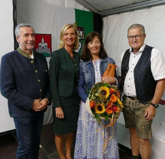 Vier Menschen, zwei Männer und zwei Frauen, stehen in einem Zelt. Eine Frau hält einen Blumenstrauß. Hinter ihnen befinden sich Banner und ein Tisch mit Flaschen und einer Rolle Papier.