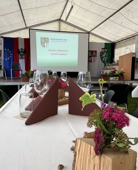 Ein Zelt-Event mit einem Tisch für eine Versammlung. Der Tisch hat Weingläser, eine hölzerne Blumenarrangement und Flaggen im Hintergrund. Eine Projektionsleinwand zeigt eine Nachricht.