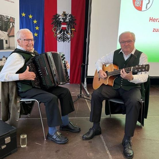 Zwei ältere Männer sitzen auf Stühlen auf einer Bühne. Einer spielt Akkordeon und der andere Gitarre. Hinter ihnen steht eine Flagge und ein Bildschirm mit deutschem Text.