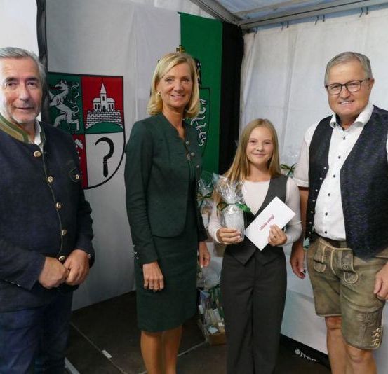 Drei Personen stehen in einem Raum, lächeln und schauen in die Kamera. Eine Frau in der Mitte hält einen Blumenstrauß und ein Zertifikat. Der Mann rechts trägt eine Uhr, und der Mann links trägt eine Brille. Hinter ihnen ist eine weiße Wand mit einer grün-roten Fahne.
