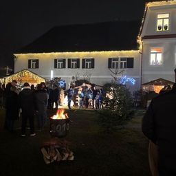 Eine Gruppe von Menschen versammelt sich um ein Lagerfeuer vor einem dekorierten Haus bei Nacht.