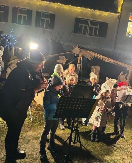 Eine Gruppe von Kindern und einem Erwachsenen spielen Flöten vor einem dekorierten Haus bei Nacht.