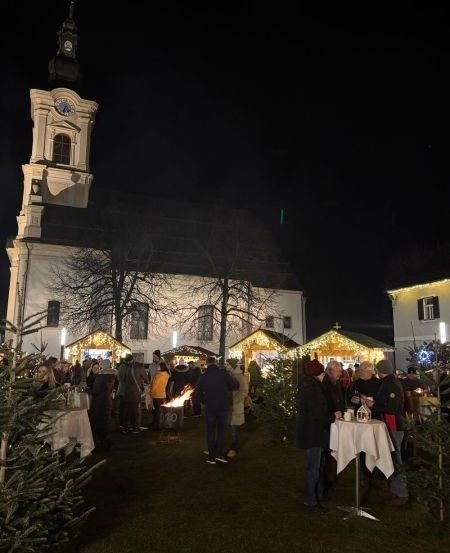 Bei Nacht versammelt sich eine Menschenmenge vor einer Kirche, mit einem Feuerkorb, Weihnachtsbäumen und festlichen Lichtern, die die Szene erhellen.