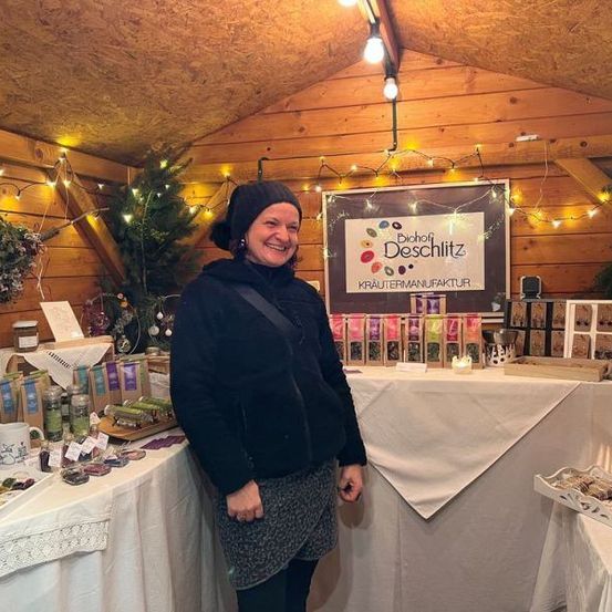 Eine Frau steht in einem Stand mit ausgestellten Produkten und lächelt. Der Stand ist mit Holzwänden, Weihnachtslichtern und Pflanzen dekoriert. Es gibt ein Schild mit dem Text Biohof Deschiitz und einem Logo.