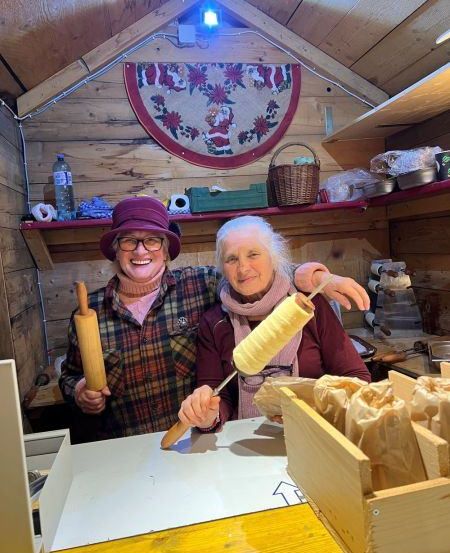 Zwei Frauen stehen in einem kleinen Holzladen und halten Holz-Nudelholz. Eine Frau lächelt, und die andere posiert mit einer Spule Garn. Dahinter befindet sich ein Regal mit einem Korb und einem Kranz.