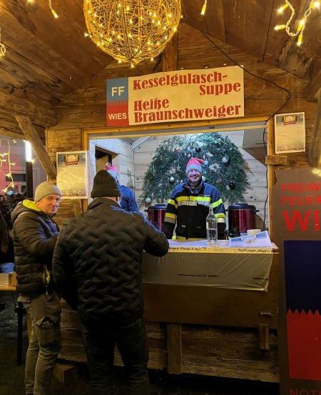 Zwei Männer stehen lächelnd an einem Essensstand, auf dem ein Schild mit der Aufschrift 'Kesselgulasch-Suppe Heiße Braunschweiger' steht. Der Stand hat einen Tisch mit einer Flasche und einem roten Behälter. Hinter dem Stand ist ein Weihnachtsbaum zu sehen.