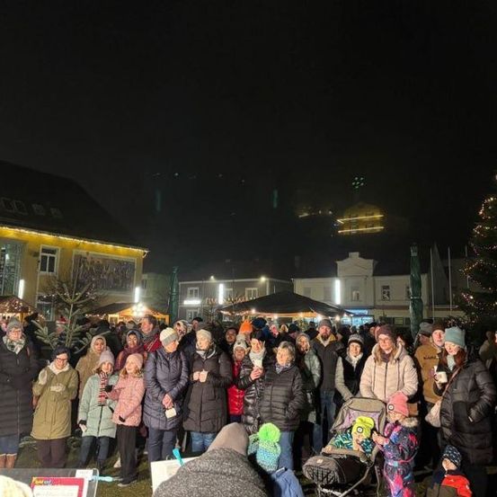 Eine Menschenmenge versammelt sich bei Nacht draußen in der Nähe eines geschmückten Weihnachtsbaums. Einige tragen Winterkleidung, Mützen und Handschuhe. Einige halten Tassen und andere machen Fotos. Gebäude mit Lichtern sind im Hintergrund.