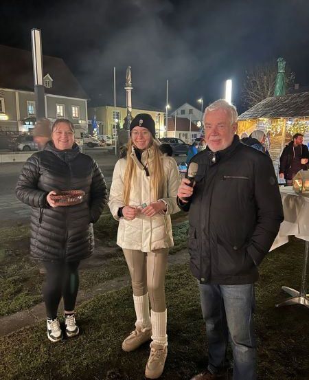 Drei Menschen stehen bei Nacht auf einem Rasen. Der Mann hält ein Mikrofon und spricht zu den beiden Frauen. Die Frau auf der linken Seite hält eine braune Schüssel. Die Frau in der Mitte trägt einen weißen Mantel. Dahinter befindet sich ein dekorierter Bereich mit einem Tisch und einem Zelt.