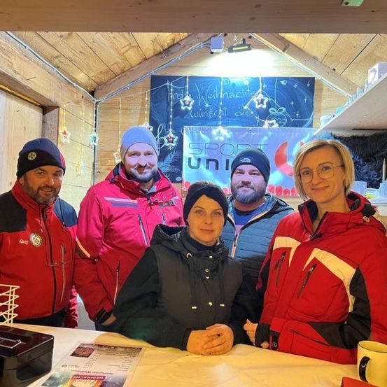 Eine Gruppe von fünf Personen in Winterjacken und Mützen posiert für ein Foto in einer Holzhütte mit einem Banner an der Wand, auf dem 'SPORT UNI' steht.