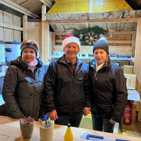 Drei Menschen stehen in einer Hütte, alle tragen Winterkleidung. Zwei Frauen und ein Mann lächeln für ein Foto. Ein Feuerlöscher und Tassen stehen auf einem Tisch vor ihnen.