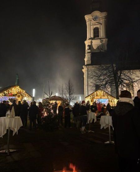 Eine Menschenmenge versammelt sich bei einer festlichen Veranstaltung im Freien, beleuchtet von Lichterketten und einem Lagerfeuer, mit einem Kirchturm im Hintergrund.