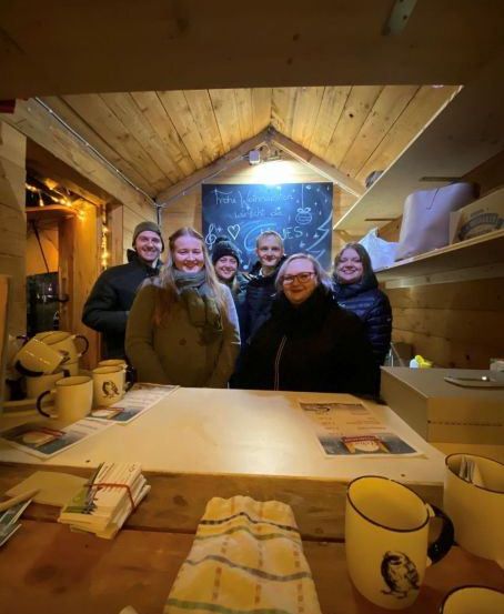 Eine Gruppe von Menschen steht in einer Holzhütte mit einem Tafelbrett hinter ihnen. Tassen, Servietten und andere Gegenstände sind auf dem Tisch.