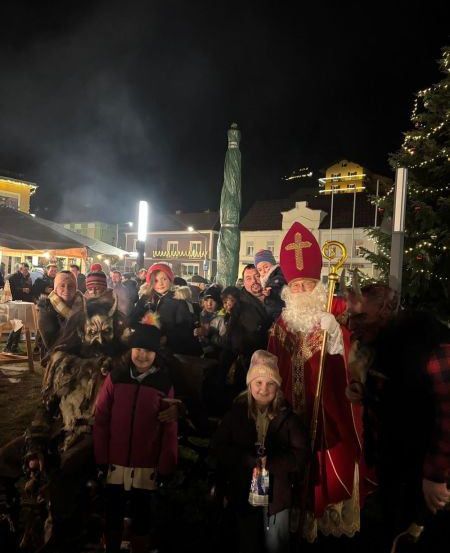 Bei Nacht versammelt sich eine Menschenmenge um einen Weihnachtsbaum und posiert mit einem als Weihnachtsmann verkleideten Mann. Dahinter befinden sich Zelte und Gebäude, und einige Leute stehen links.