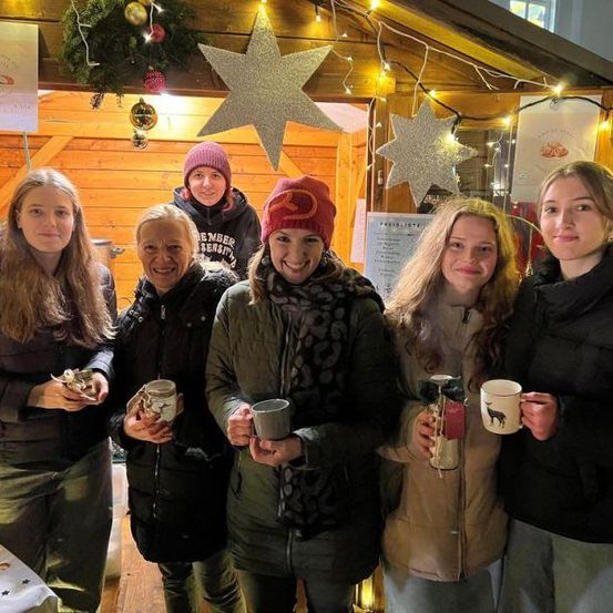 Eine Gruppe von Frauen steht lächelnd und mit Tassen in der Hand an einem Weihnachtsmarktstand. Sie tragen Wintermäntel und Schals. Hinter ihnen ist eine dekorierte Holzstruktur mit Lichtern und Ornamenten zu sehen.