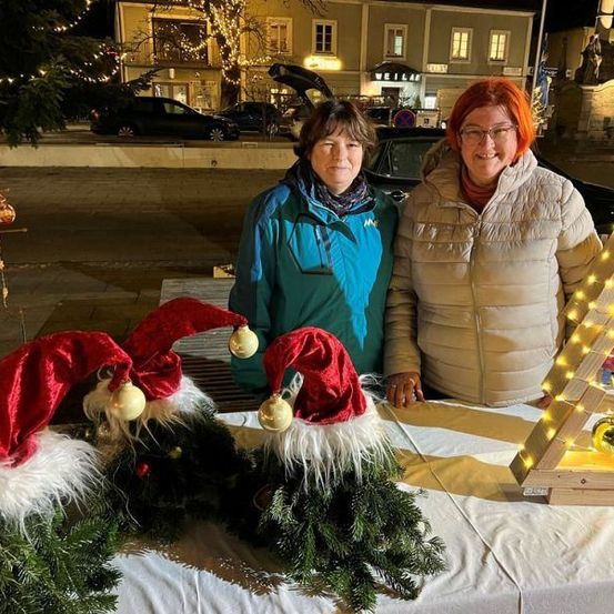 Zwei Frauen stehen hinter einem Tisch mit Weihnachtsdekoration. Eine trägt eine grüne Jacke und die andere eine beige. Dahinter steht ein mit Lichtern beleuchteter Weihnachtsbaum.