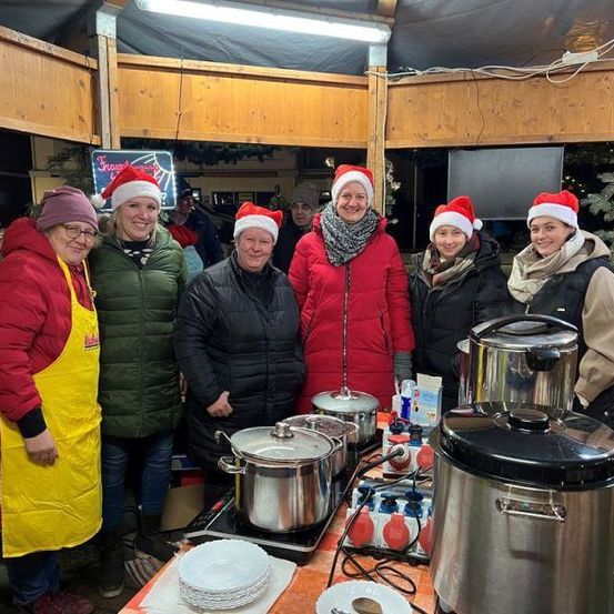 Eine Gruppe von Frauen mit Weihnachtsmützen steht nebeneinander in einer Küche. Sie lächeln und posieren für ein Foto. Auf dem Herd stehen Töpfe und Pfannen, und eine Flasche steht auf dem Tisch. Es gibt einen verschwommenen Blick auf einen Holzzaun und einen Baum draußen.
