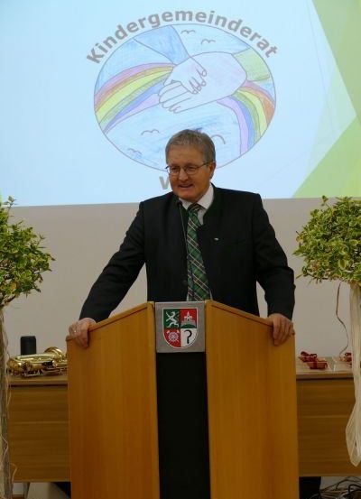 Ein Mann in formeller Kleidung steht lächelnd an einem Rednerpult. Hinter ihm befindet sich eine Projektionsleinwand mit den Worten 'Kindergemeinderat' und einem handgezeichneten Logo. Zwei Topfpflanzen flankieren das Rednerpult.