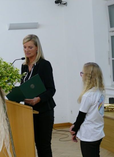 Eine Frau steht an einem Podium, hält eine grüne Mappe in der Hand und spricht in ein Mikrofon. Eine andere Frau mit Brille steht neben ihr und schaut zur Rednerin.