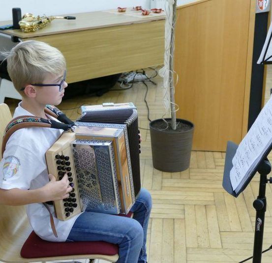 Ein junger Junge in einem weißen Hemd und einer Brille spielt ein Akkordeon, während er auf einem Stuhl sitzt. Ein Notenständer mit einem Notenblatt steht in der Nähe, und eine Saxophon befindet sich auf dem Schreibtisch hinter ihm.